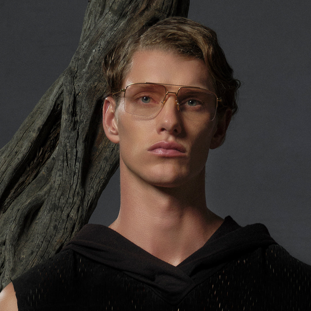 A person with short hair and gold-rimmed glasses gazes forward. They wear a black top, standing beside a textured tree trunk, set against a dark background.