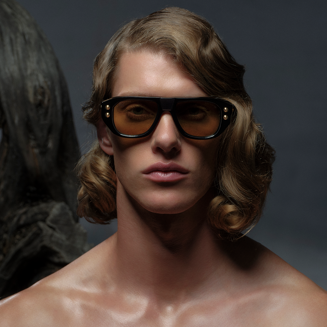 A person with wavy, shoulder-length hair wears oversized, brown sunglasses. The composition is dramatic, with neutral tones and a shadowed background, conveying a modern, stylish vibe.