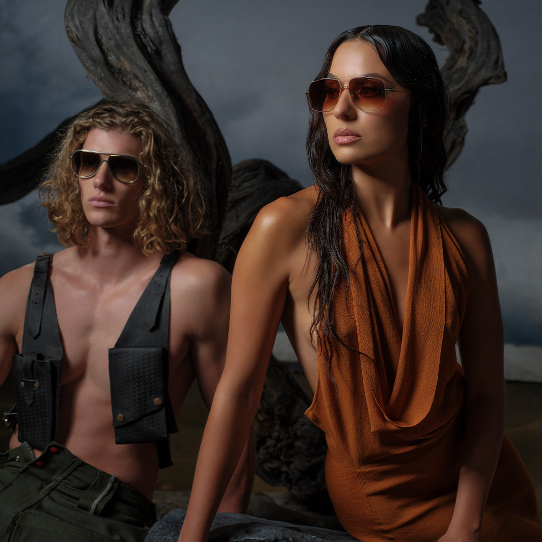Two models with long, wavy hair wearing stylish sunglasses pose against a dramatic, stormy sky and twisted wood backdrop. The scene is intense and fashion-forward.