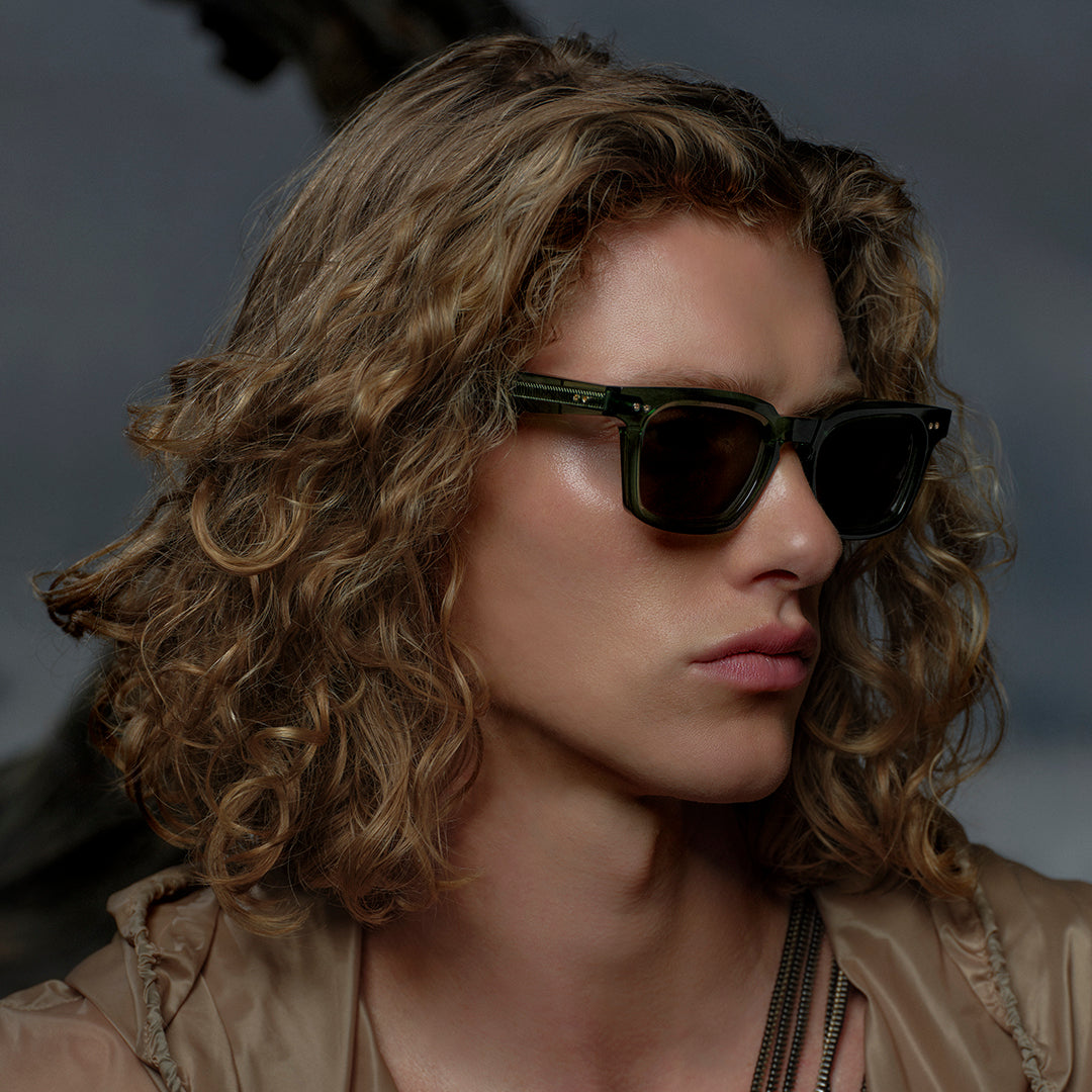 Person with curly blonde hair wearing dark sunglasses, looking sideways. The background is blurred, conveying a mysterious and stylish tone.