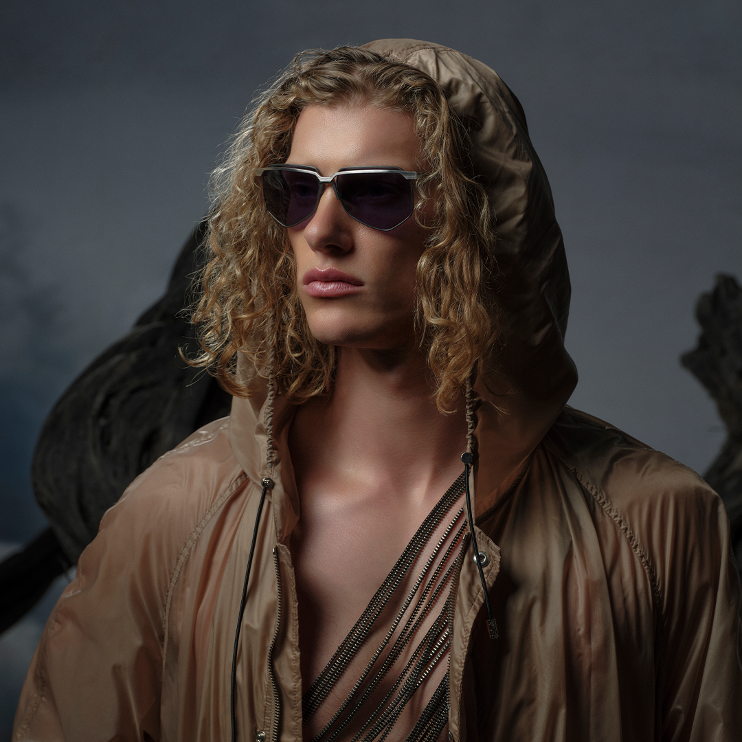 Young person with curly hair and sunglasses wears a tan hooded jacket in a moody setting. The expression is serious, conveying a sense of mystery.