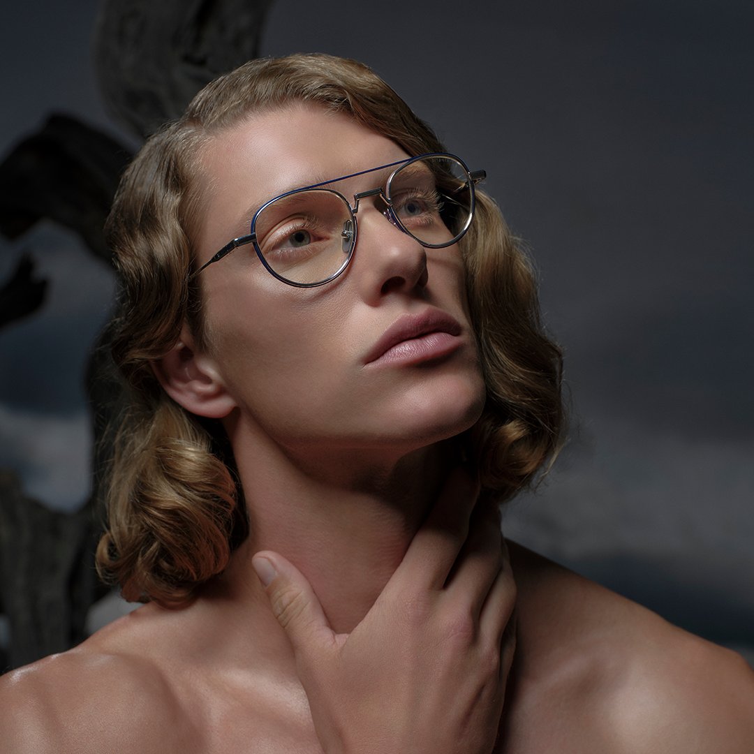 A person with wavy hair and glasses gazes upward thoughtfully, hand touching their neck. The background is dark and moody, suggesting contemplation.