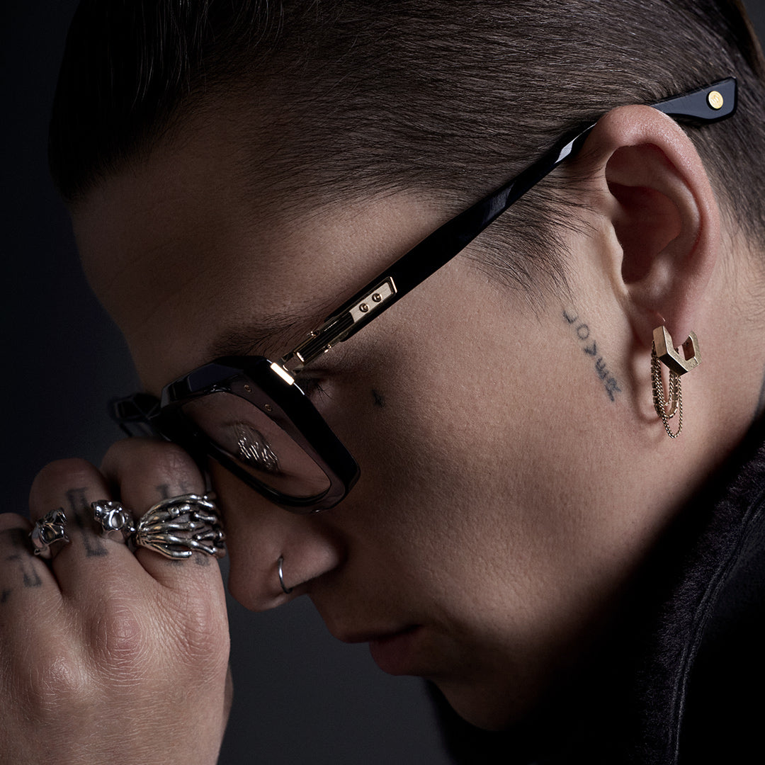 A person with wavy hair and glasses gazes upward thoughtfully, hand touching their neck. The background is dark and moody, suggesting contemplation.