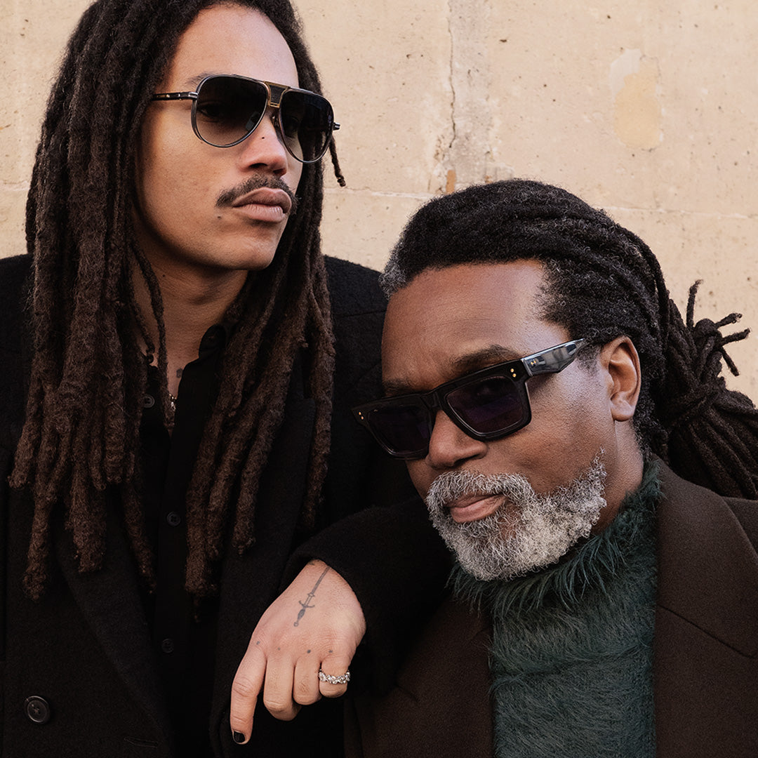 Two men with dreadlocks and sunglasses, dressed in stylish dark clothing, pose confidently against a beige wall, exuding a cool, composed vibe.
