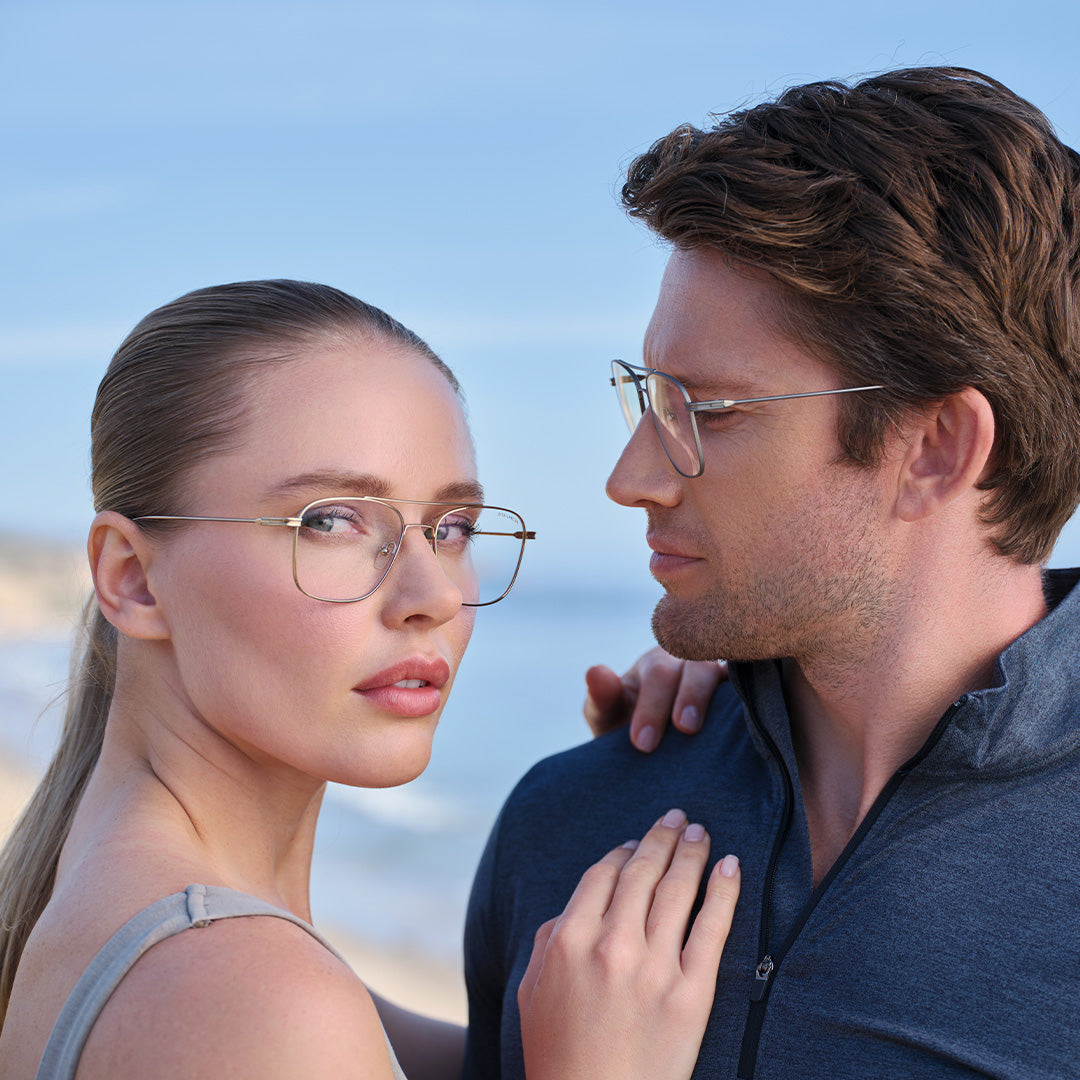A woman and a man wearing glasses gaze at each other near a beach. The sky is clear, conveying a serene and intimate mood. The woman looks directly at the viewer.