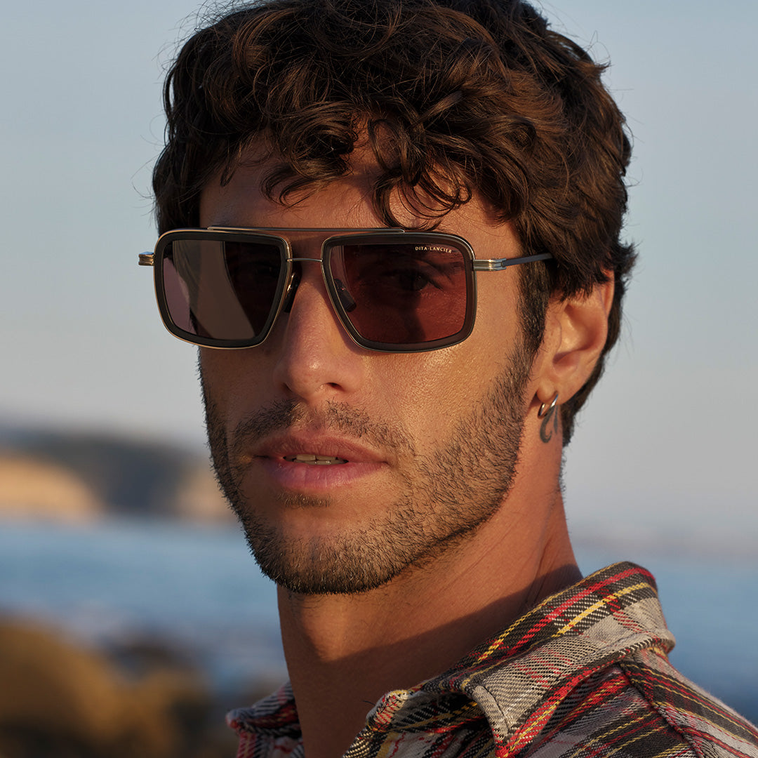 A man with curly hair and a short beard wearing square sunglasses gazes into the distance by the sea. He's dressed in a plaid shirt, evoking a relaxed, coastal vibe.