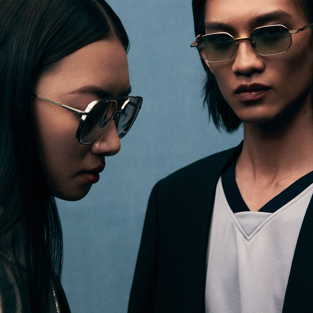 Two people wearing sunglasses stand closely against a blue background. The mood is serious and stylish, with a focus on fashion and modern aesthetics.