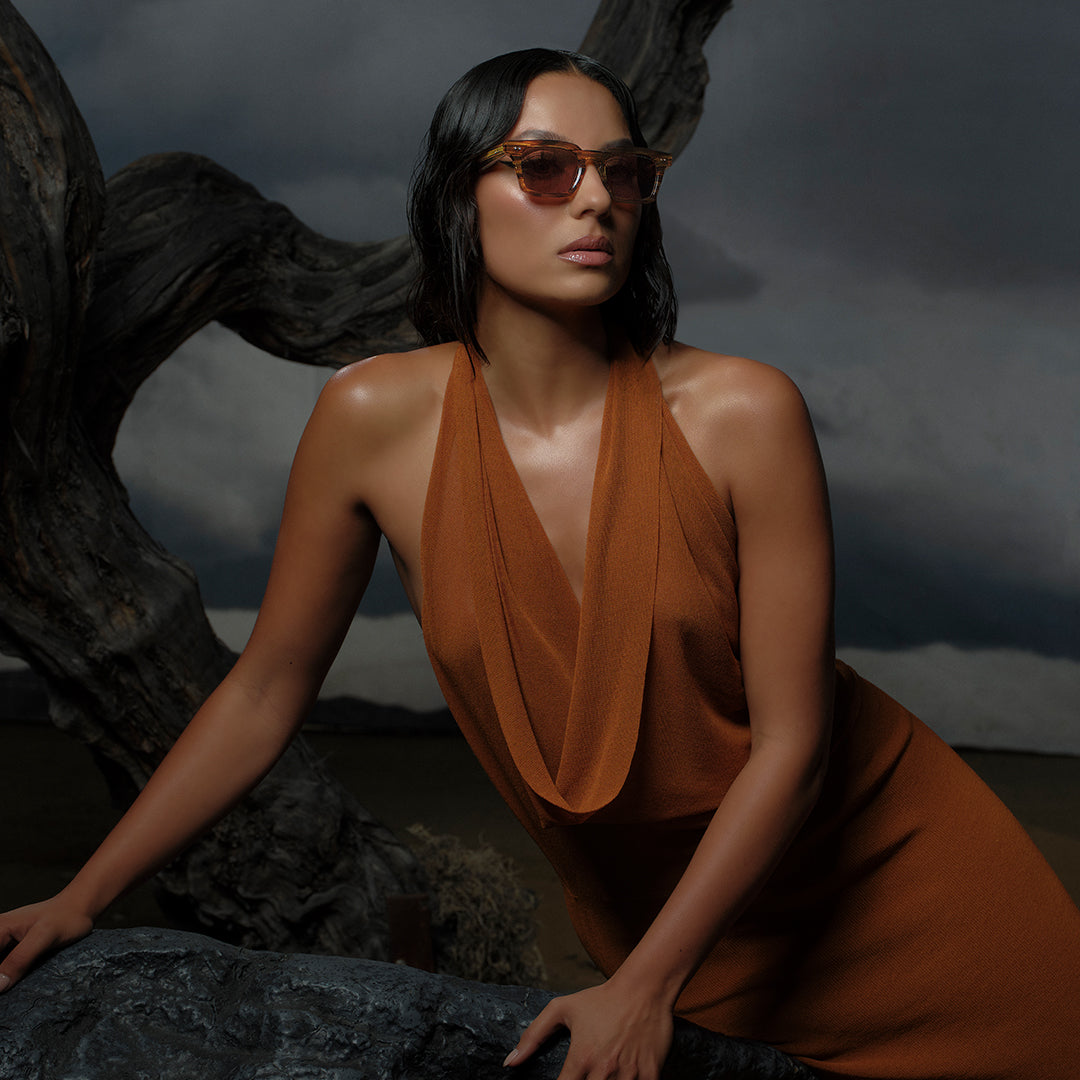 A woman in a rust-colored dress and sunglasses leans against a stone under a dark, cloudy sky. Her pose is confident, with a dramatic, moody tone.