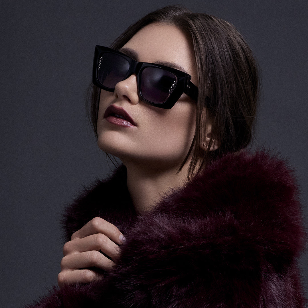 A woman in a rust-colored dress and sunglasses leans against a stone under a dark, cloudy sky. Her pose is confident, with a dramatic, moody tone.