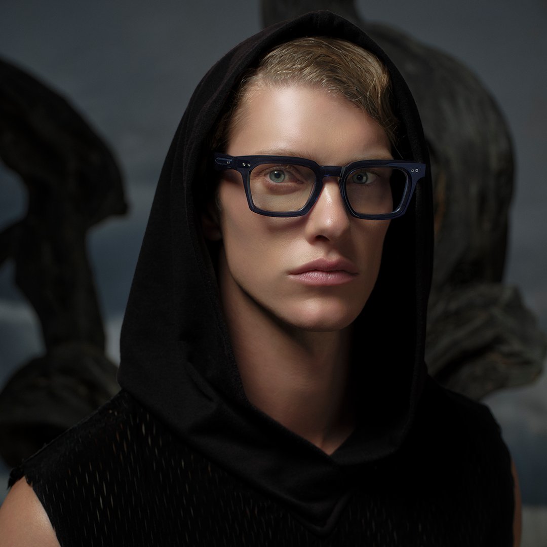 Person with short hair, wearing a black hood and glasses, looks directly ahead with a neutral expression. Dark, moody background adds intensity.