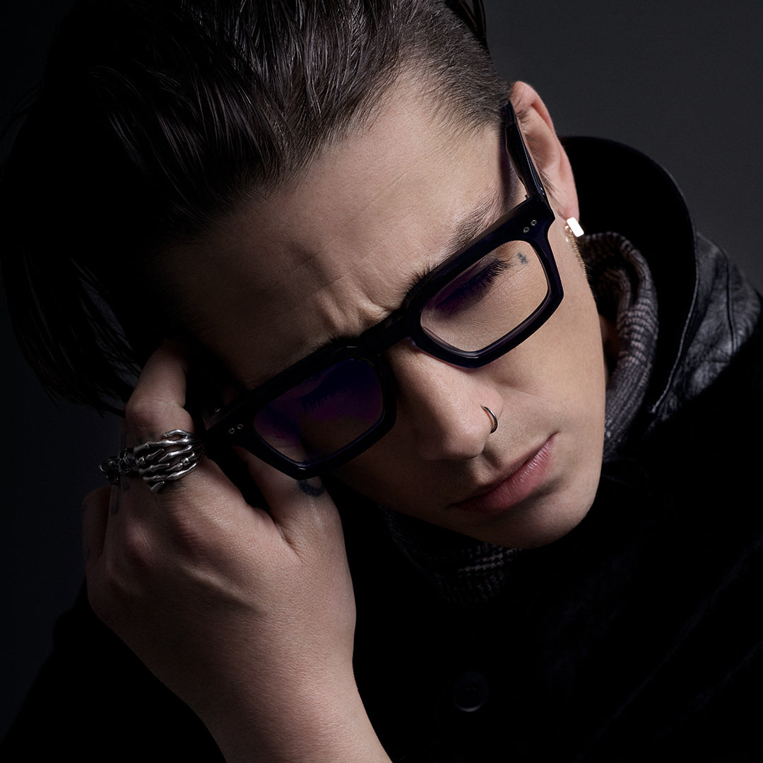 Person with short hair, wearing a black hood and glasses, looks directly ahead with a neutral expression. Dark, moody background adds intensity.