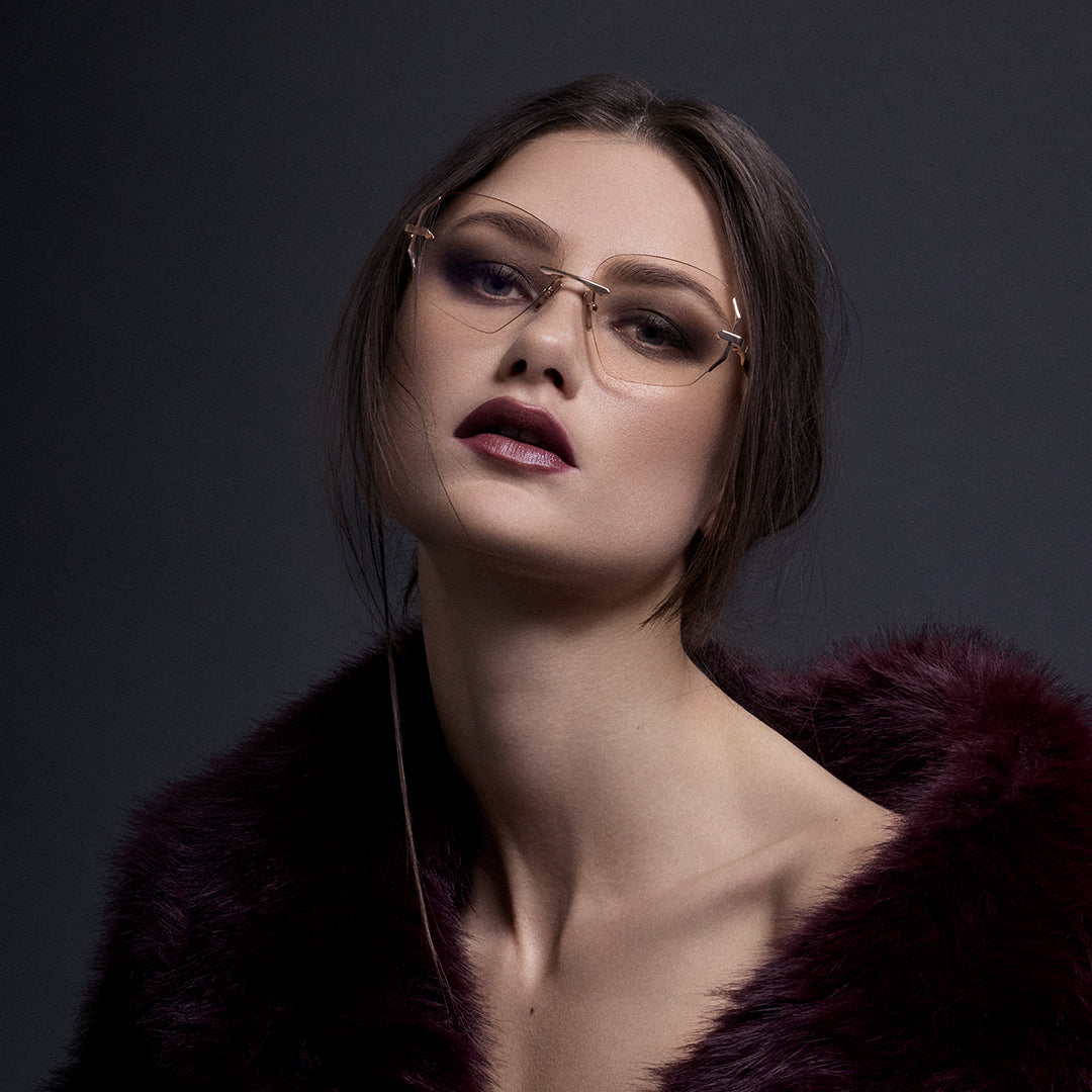 A woman with long blonde hair sits against twisted driftwood, wearing a bare-back top with glittering details and a flowing white skirt, in a moody, overcast setting.