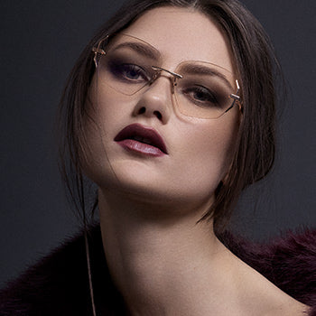 A woman with long blonde hair sits against twisted driftwood, wearing a bare-back top with glittering details and a flowing white skirt, in a moody, overcast setting.