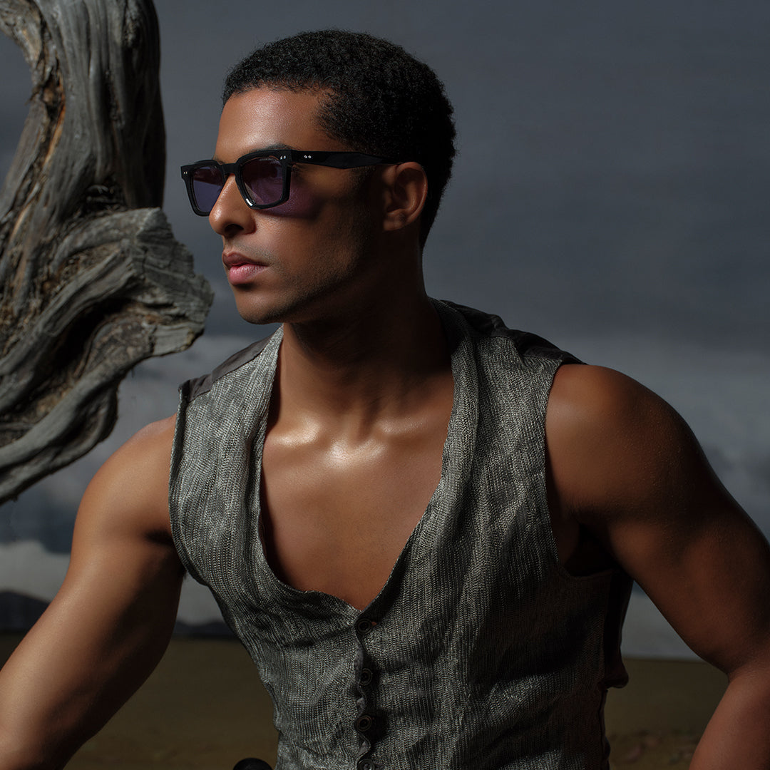 Young man with short hair wearing sunglasses and a sleeveless gray vest, standing outdoors against a dramatic, clouded sky and a twisted tree.