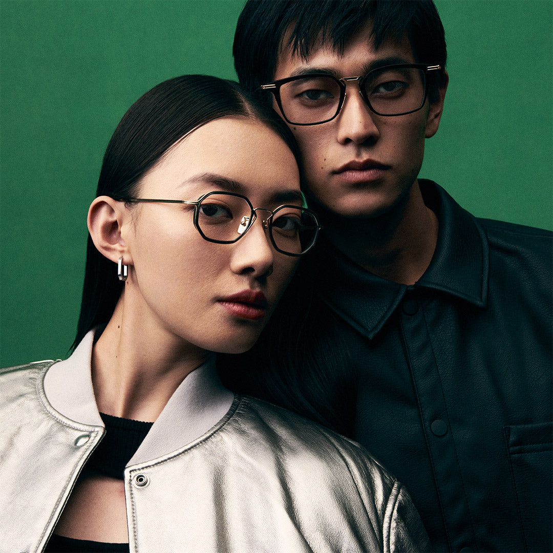 A woman in a silver jacket and a man in a dark leather shirt pose against a green background. Both wear stylish glasses, looking confident and composed.