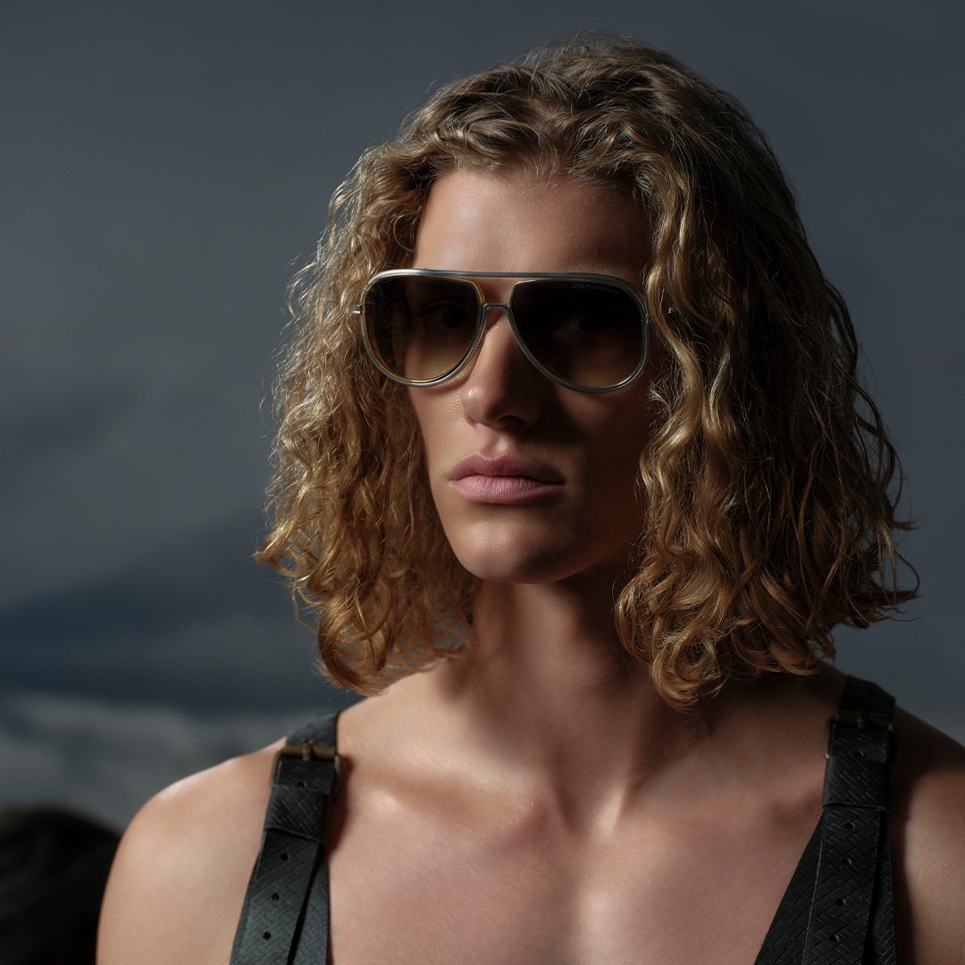 A person with long, curly hair wears aviator sunglasses and a dark strapped top against a moody, cloudy backdrop, conveying a confident, edgy vibe.