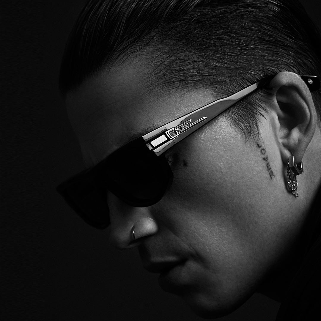 A person with long, curly hair wears aviator sunglasses and a dark strapped top against a moody, cloudy backdrop, conveying a confident, edgy vibe.