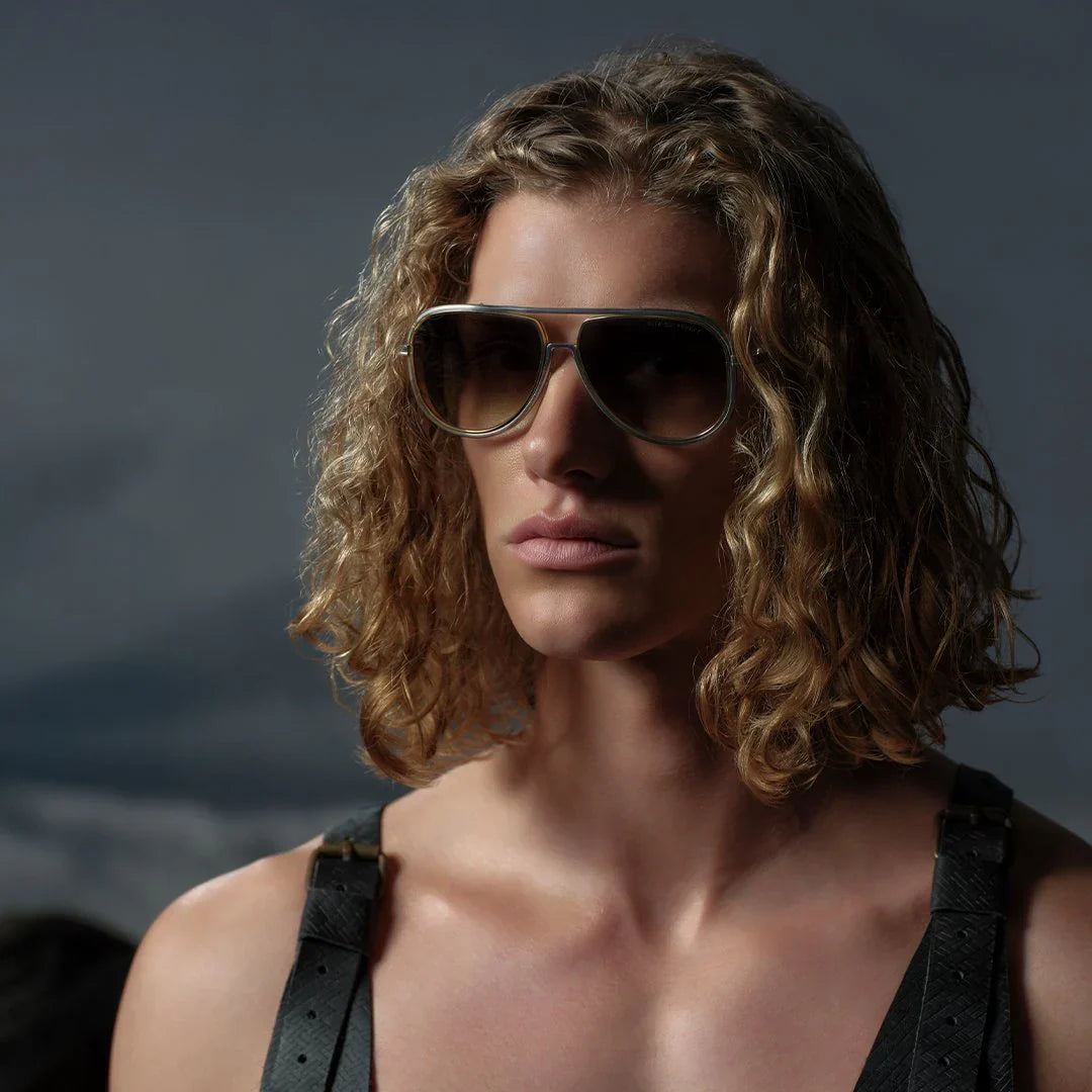 Young person with long curly hair and sunglasses, wearing a leather strap over one shoulder, against a dark, moody sky, exuding a confident and edgy vibe.