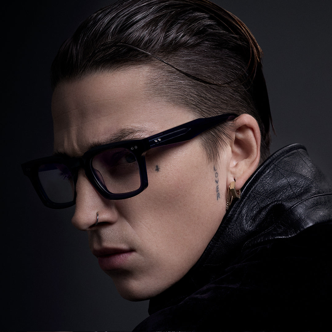 A person with short hair and gold-rimmed glasses gazes forward. They wear a black top, standing beside a textured tree trunk, set against a dark background.