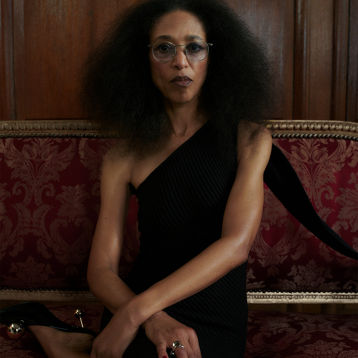 A person with short hair and gold-rimmed glasses gazes forward. They wear a black top, standing beside a textured tree trunk, set against a dark background.