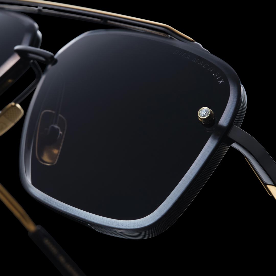 Close-up of a luxurious pair of sunglasses with sleek, dark lenses and a metallic frame. A small diamond accent sparkles on the frame's corner. Elegant design.