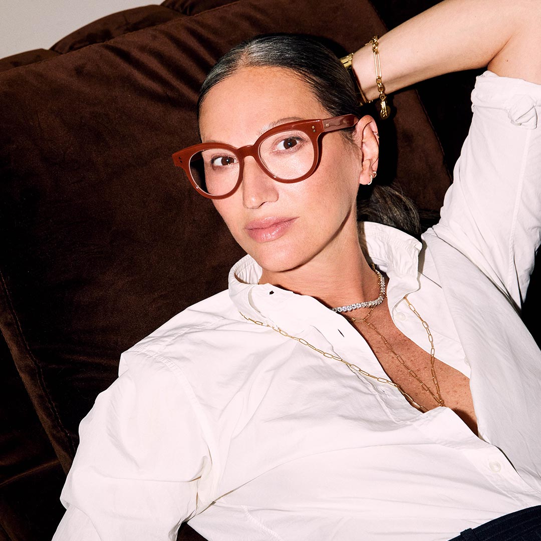 A person with brown glasses and slicked-back hair lounges on a dark sofa. They wear a white shirt, layered necklaces, and exude a relaxed, confident vibe.