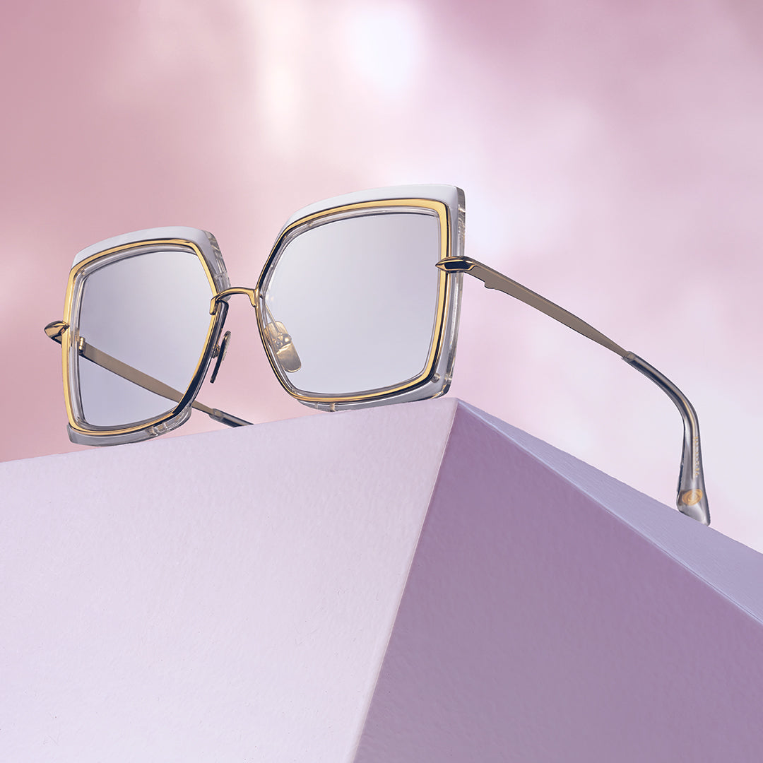 Square-framed eyeglasses with thin gold rims rest on a geometric lavender block, set against a soft pink, dreamy background.