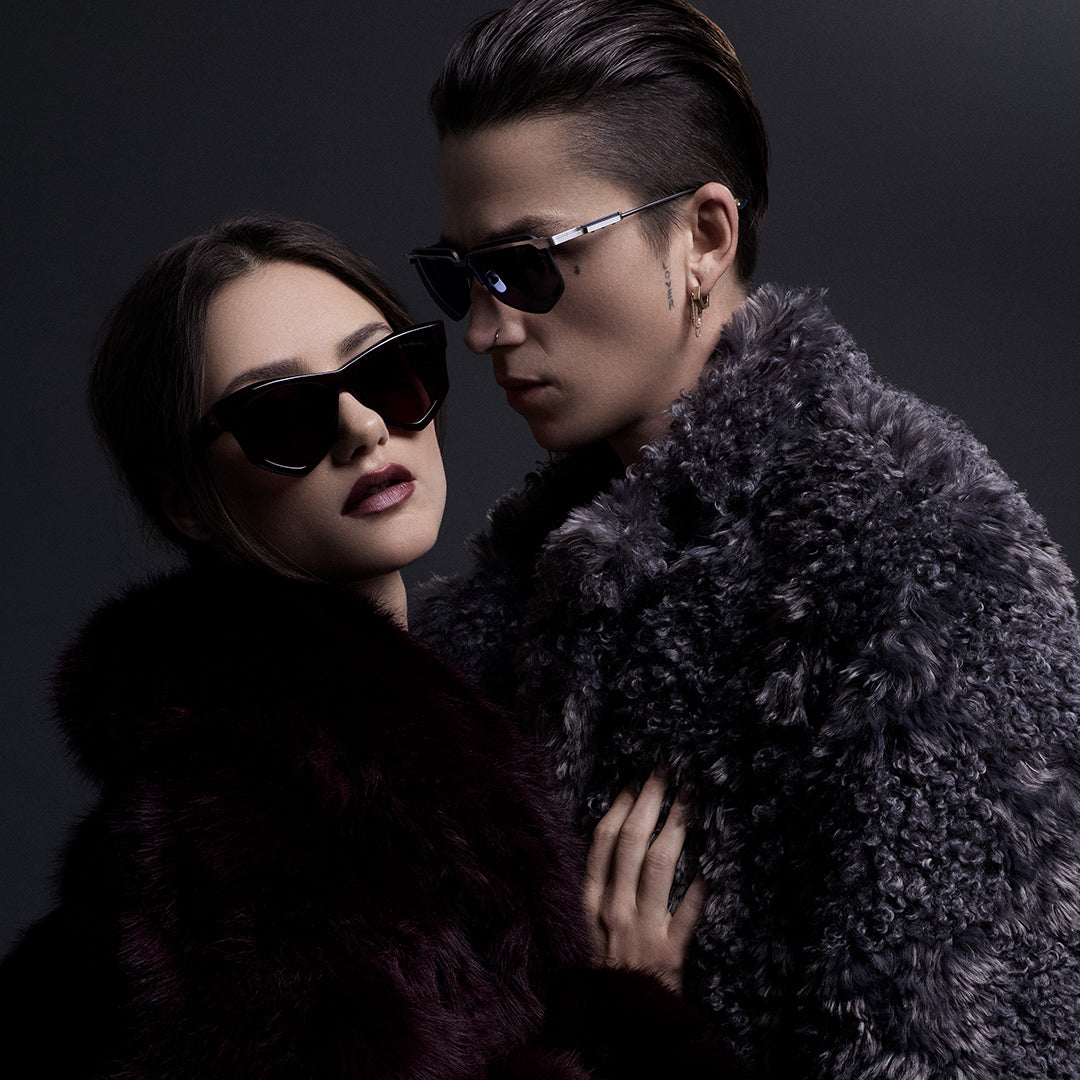 Two individuals in stylish, modern outfits stand in an arid landscape. One is seated on a rock; the other stands, both wearing glasses, under moody skies.