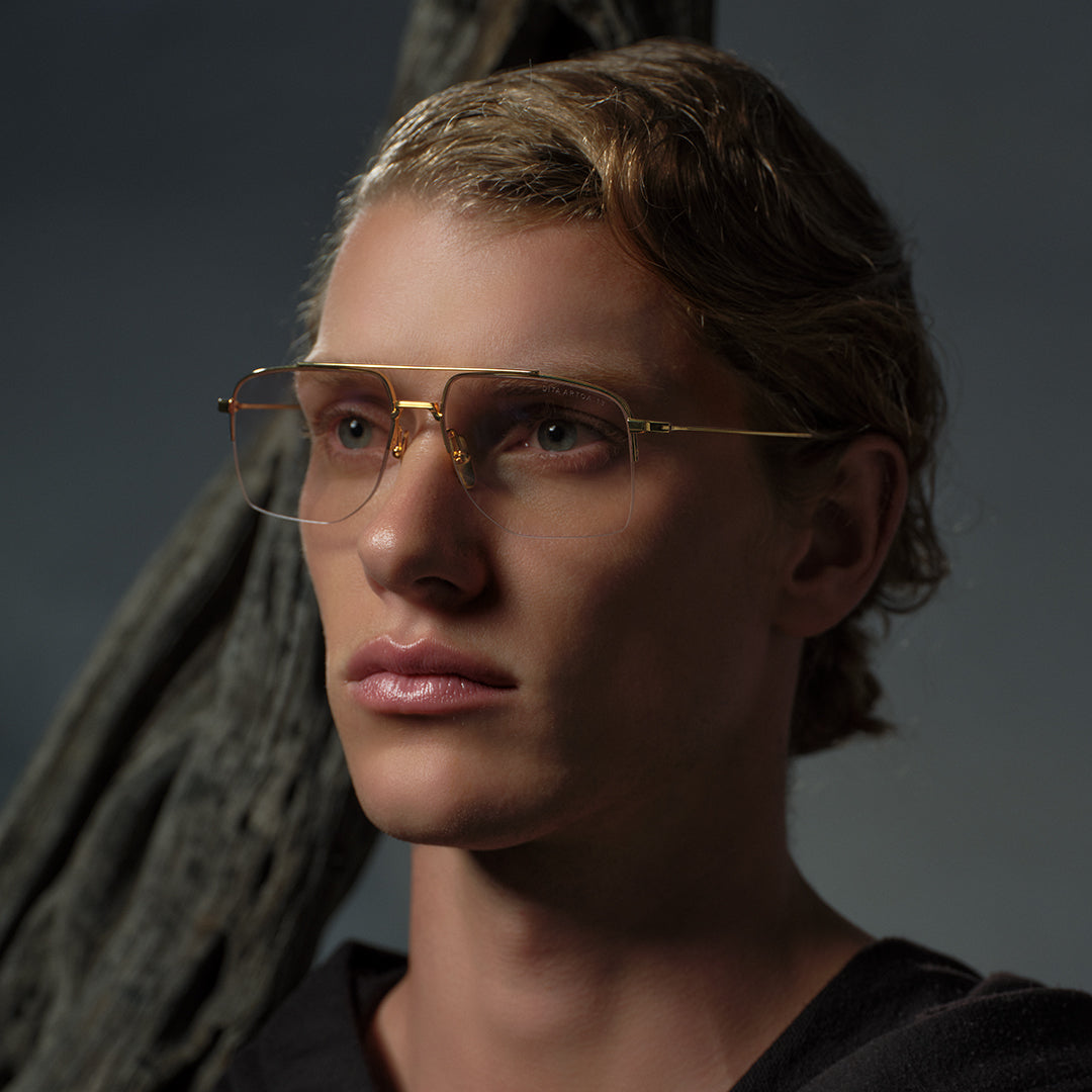 A person with wavy blonde hair and gold-rimmed glasses gazes thoughtfully into the distance against a dark, textured background. The mood is contemplative.