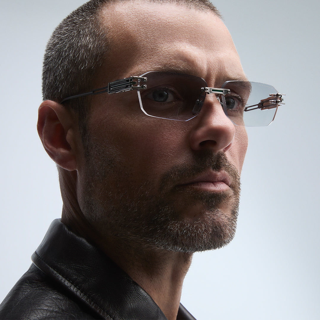 A person with wavy blonde hair and gold-rimmed glasses gazes thoughtfully into the distance against a dark, textured background. The mood is contemplative.