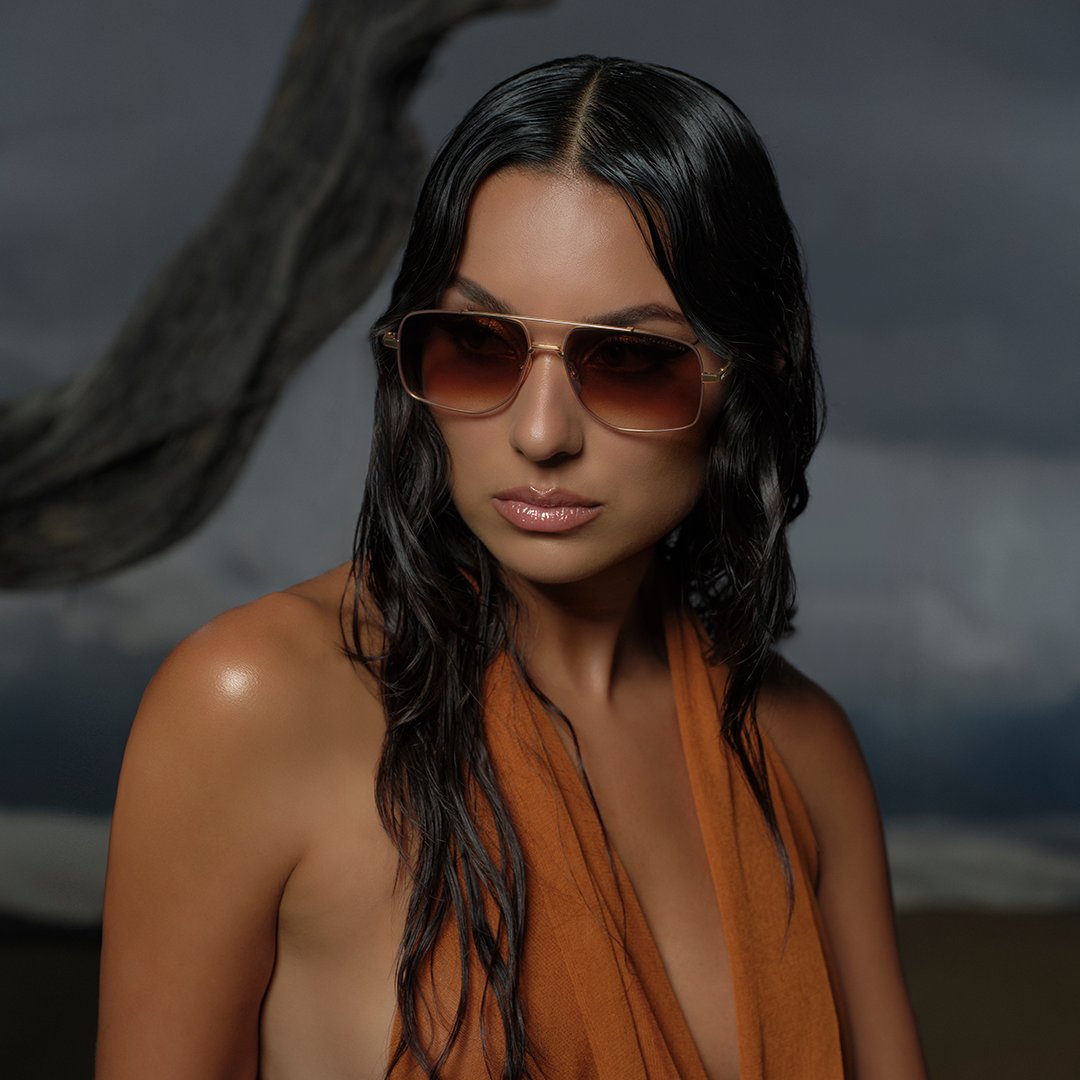 A woman with long dark hair and wet skin wears large brown sunglasses and an orange halter top. She stands against a moody, cloudy backdrop, evoking a dramatic and stylish tone.