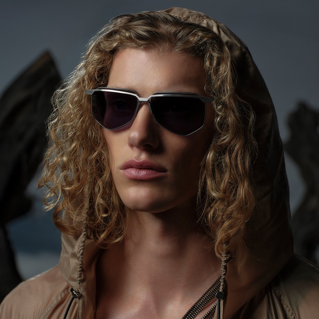Young adult with curly hair, wearing dark sunglasses and a hooded jacket, poses against a moody background, conveying a cool, mysterious vibe.