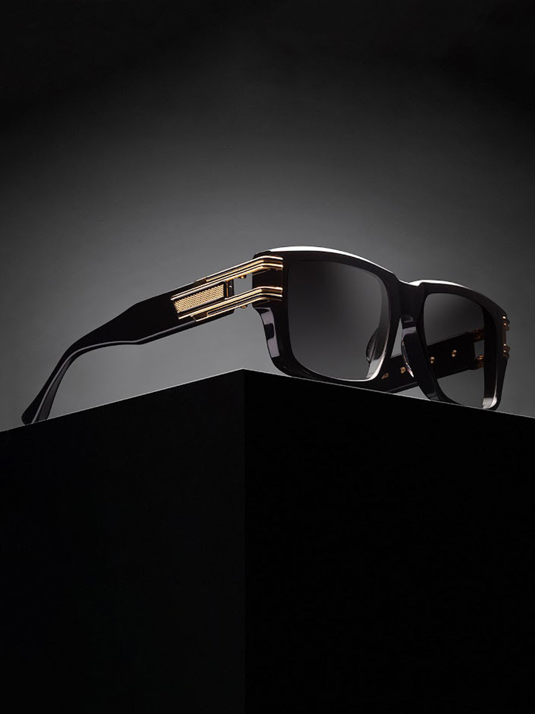 Sleek black sunglasses with gold accents rest on a dark pedestal against a moody gradient background, conveying elegance and luxury.