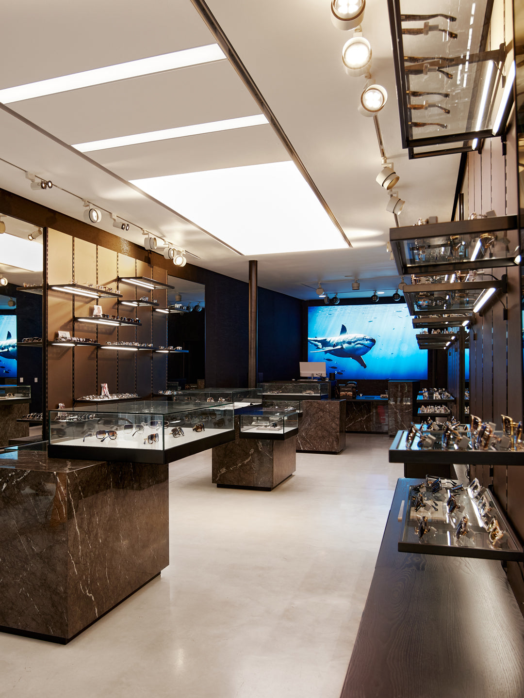 Elegant eyewear store interior with sleek glass display cases and marble accents; a large screen shows a swimming shark, adding a modern touch.