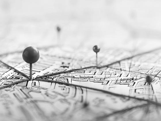 A black and white image shows pushpins on a detailed map, connected by lines. The scene suggests planning or travel with a focused, strategic tone.