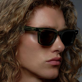 A person with curly hair wearing dark sunglasses gazes sideways against a moody, dark background. The tone is calm and introspective.