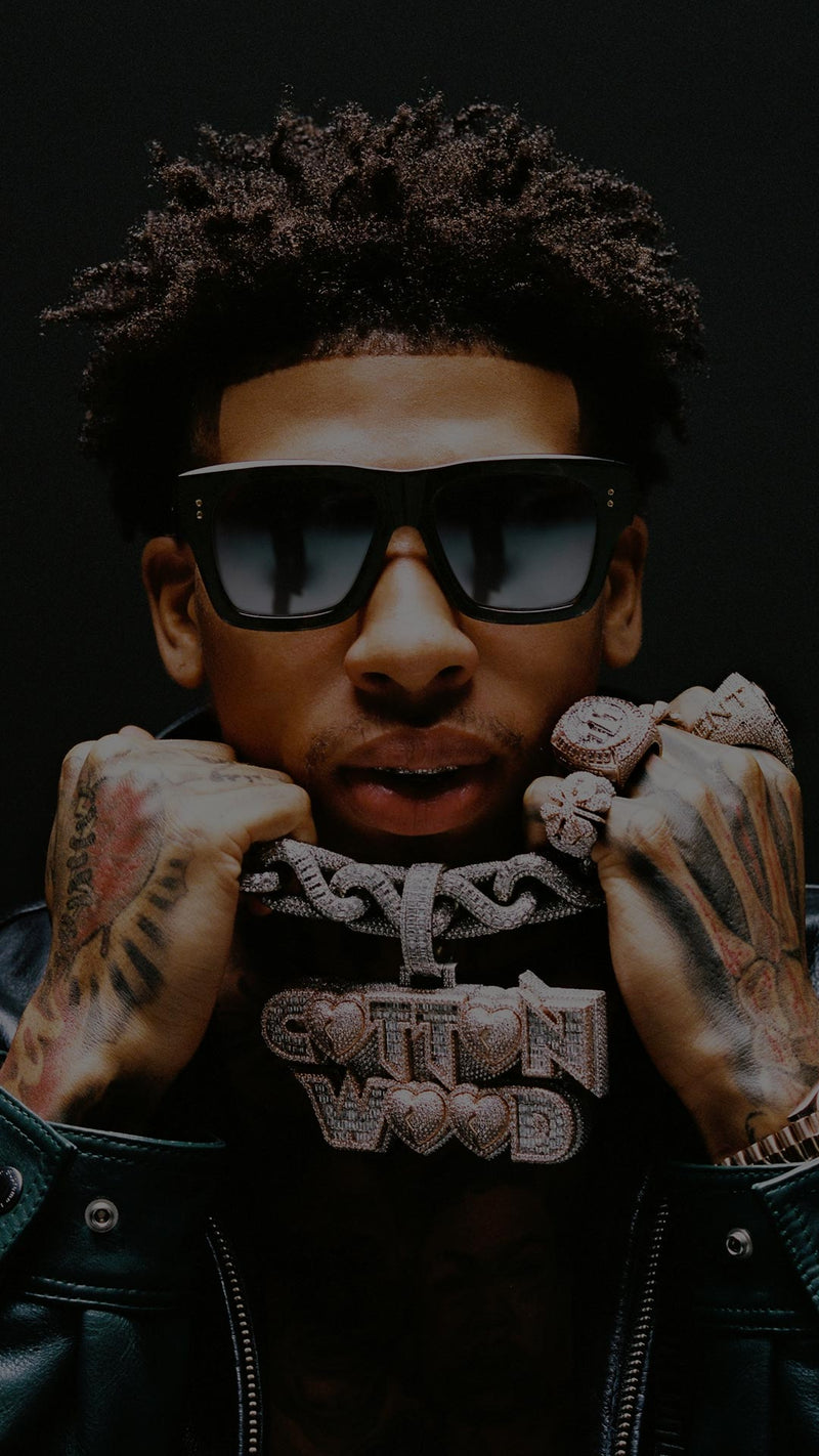 NLE Choppa with curly hair wearing sunglasses and a leather jacket displays a large, ornate chain necklace. His hands, decorated with rings, hold the chain confidently.
