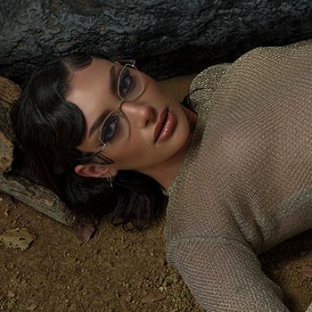 Woman with short curly hair in glasses, wearing a glittery mesh top, lies on sandy ground with a contemplative expression. Earthy tones prevail.