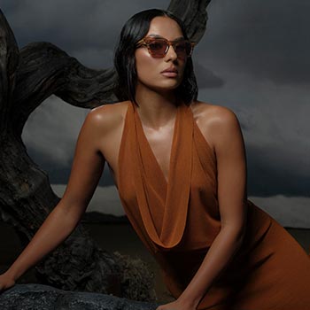 A woman with short, dark hair wearing sunglasses and a rust-colored halter dress leans on a textured rock. The background features dramatic, cloudy skies.
