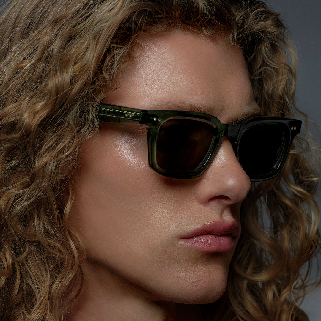 A person with long curly hair wearing sleek, dark sunglasses looks to the side against a muted background. The expression is confident and calm.