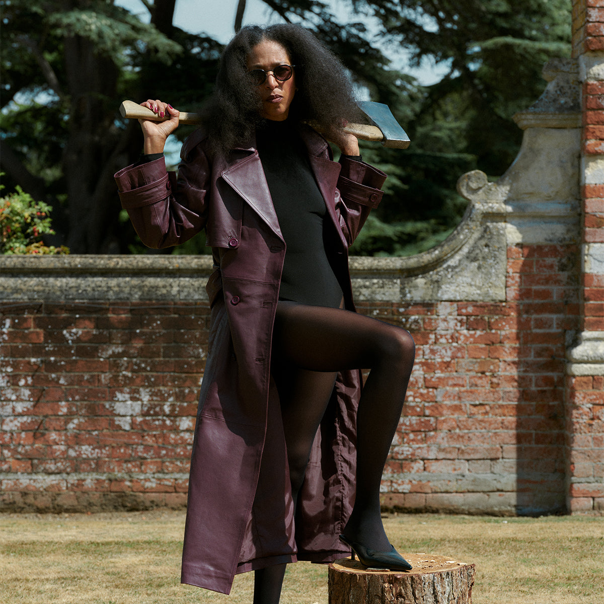 A woman in a rust-colored dress and sunglasses leans against a stone under a dark, cloudy sky. Her pose is confident, with a dramatic, moody tone.