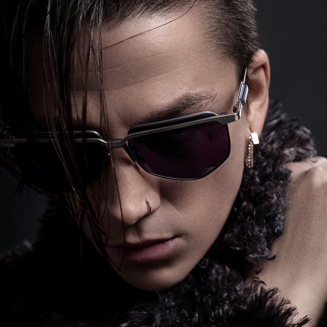 Young person with long curly hair and sunglasses, wearing a leather strap over one shoulder, against a dark, moody sky, exuding a confident and edgy vibe.