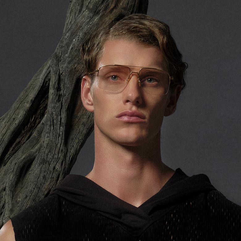 A person with short wavy hair wears gold-framed glasses and a black textured top, standing in front of a large, twisted tree branch, conveying a calm, modern tone.