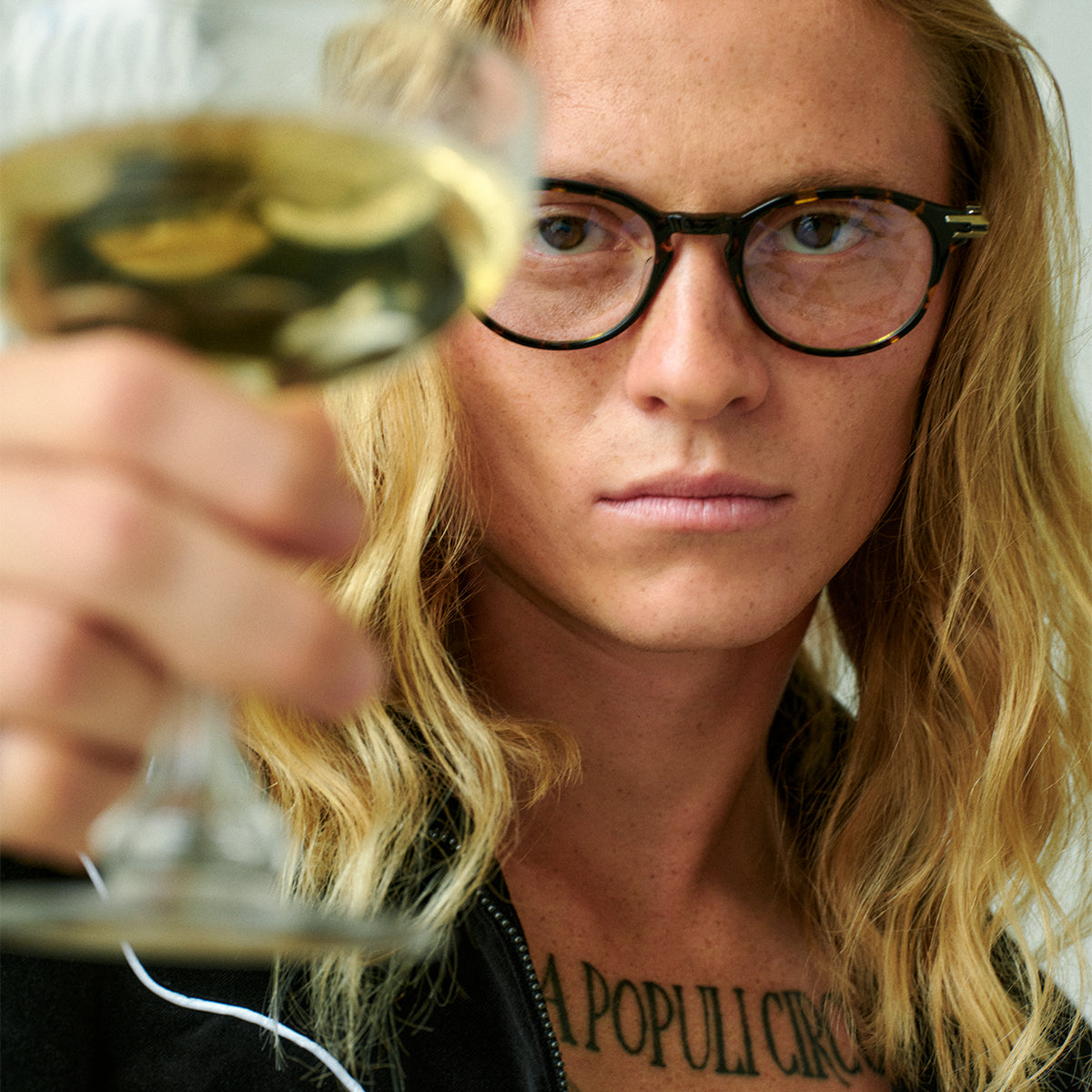 A person with wavy blonde hair and gold-rimmed glasses gazes thoughtfully into the distance against a dark, textured background. The mood is contemplative.