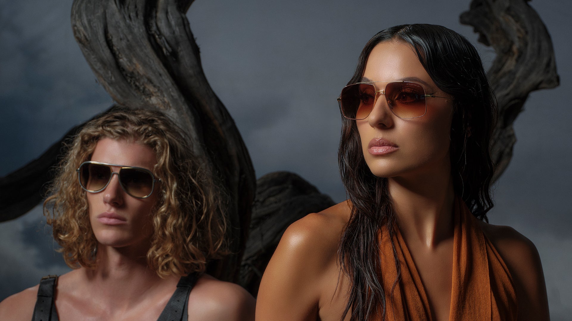 Two models in sunglasses pose confidently in a dramatic, moody setting with twisted driftwood. They wear stylish outfits in earthy tones, exuding a bold and fashionable vibe.