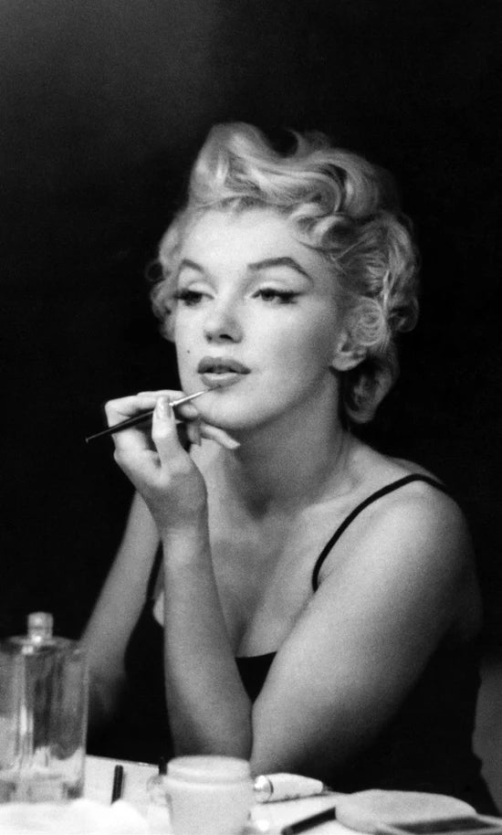 Marilyn with blonde curls applies lipstick while looking into a mirror. She wears a black tank top, exuding a focused and glamorous mood.
