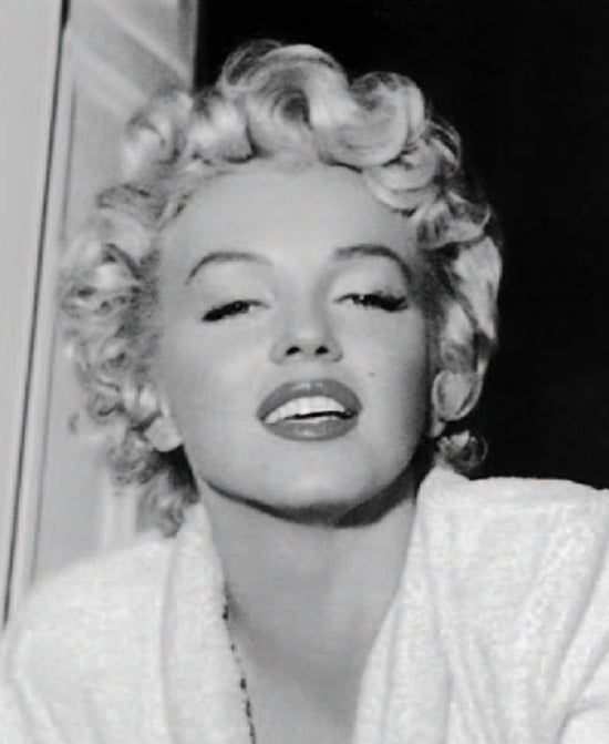 Marilyn Monroe with curly blonde hair and a white robe gazes upward with a confident expression. The image has a classic, glamorous feel.
