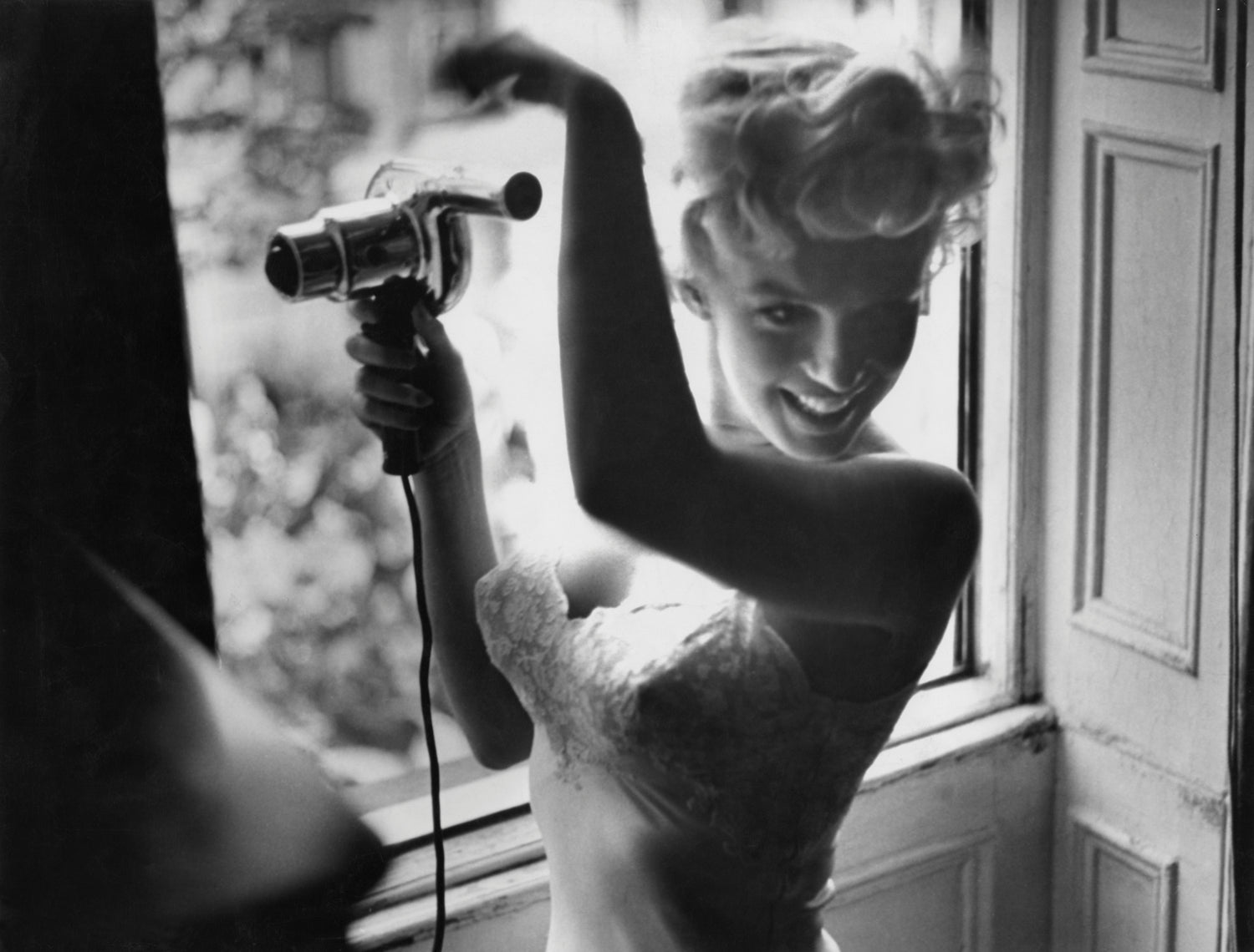 Marilyn Monroe with curled hair uses a hairdryer by a sunlit window, wearing a strapless top. She smiles, creating a playful and cheerful mood.